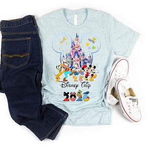 May include: A light blue heathered t-shirt featuring a graphic of a castle, Disney characters, the text Disney Trip, and the year 2026, paired with dark blue denim jeans and white canvas sneakers. The graphic includes a Mickey Mouse silhouette.
