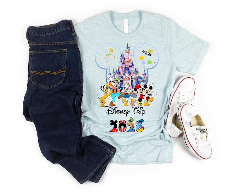 May include: A light blue heathered t-shirt featuring a graphic of a castle, Disney characters, the text Disney Trip, and the year 2026, paired with dark blue denim jeans and white canvas sneakers. The graphic includes a Mickey Mouse silhouette.