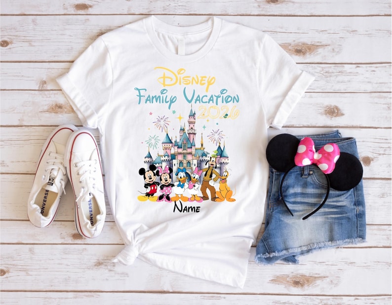 May include: A white t-shirt featuring a colorful graphic of a castle, fireworks, and Disney characters including Mickey, Minnie, Donald, Daisy, Goofy, and Pluto. The text reads Disney Family Vacation 2026 and Name. White sneakers and denim shorts with mouse ears are also shown.