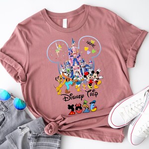 May include: A mauve short-sleeve t-shirt featuring a graphic of a castle inside a Mickey Mouse silhouette outline, surrounded by Disney characters, the text "NAME", "Disney Trip", and "2026" with character-themed numbers.
