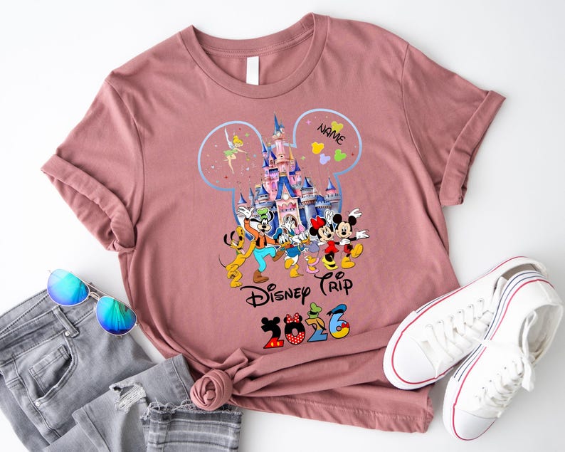 May include: A mauve short-sleeve t-shirt featuring a graphic of a castle inside a Mickey Mouse silhouette outline, surrounded by Disney characters, the text "NAME", "Disney Trip", and "2026" with character-themed numbers.