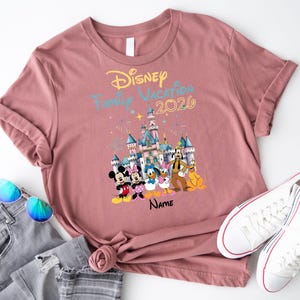 May include: A mauve short-sleeved t-shirt featuring a graphic of a castle, fireworks, and Disney characters including Mickey, Minnie, Donald, Daisy, Goofy, and Pluto. The text reads Disney Family Vacation 2026 and Name in black script.