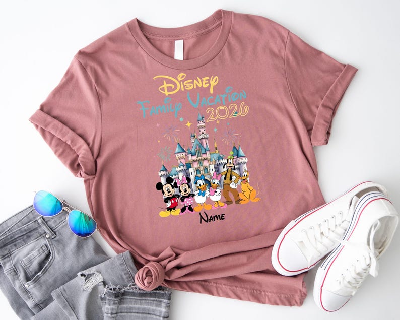May include: A mauve short-sleeved t-shirt featuring a graphic of a castle, fireworks, and Disney characters including Mickey, Minnie, Donald, Daisy, Goofy, and Pluto. The text reads Disney Family Vacation 2026 and Name in black script.