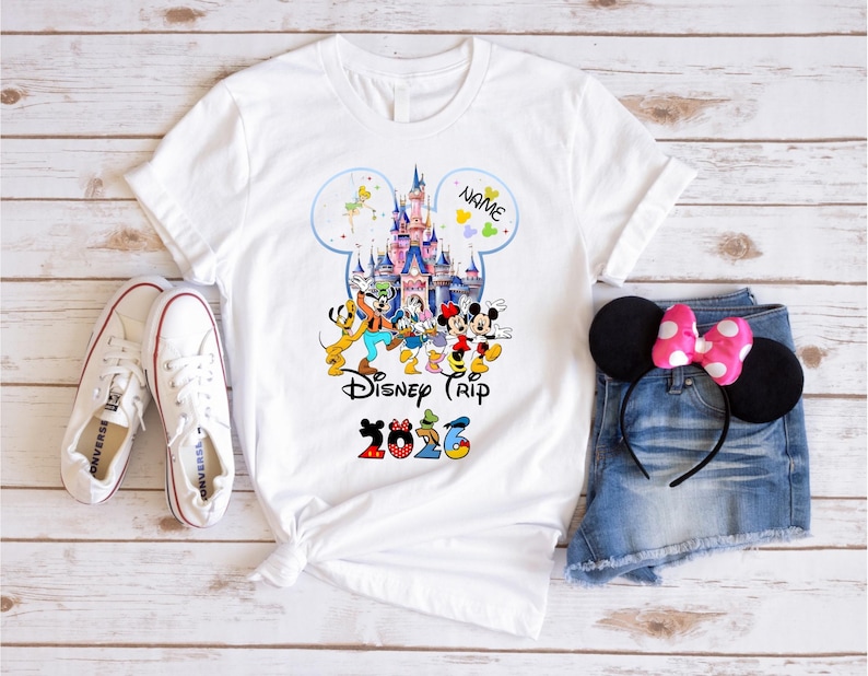 May include: A white t-shirt featuring a colorful graphic of a castle and various cartoon characters, with the text "NAME", "Disney Trip", and "2026" printed on the front. The shirt is displayed alongside white sneakers and denim shorts.