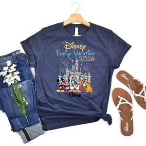 May include: A navy blue short-sleeve t-shirt featuring a graphic of Disney characters in front of a castle with fireworks. The text reads Disney Family Vacation 2026 and Name. The shirt is displayed on a wooden hanger with blue jeans and sandals.