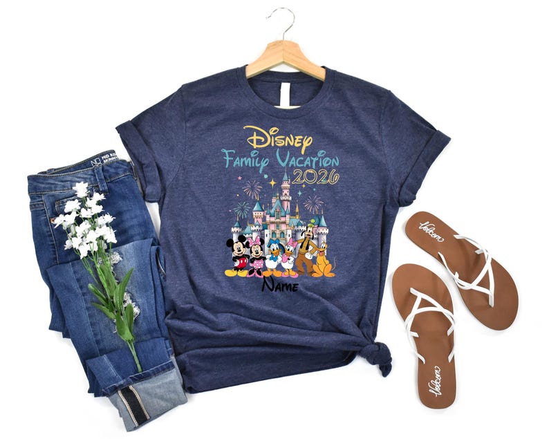 May include: A navy blue short-sleeve t-shirt featuring a graphic of Disney characters in front of a castle with fireworks. The text reads Disney Family Vacation 2026 and Name. The shirt is displayed on a wooden hanger with blue jeans and sandals.
