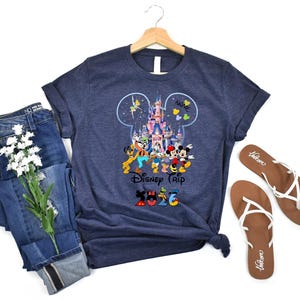 May include: A dark heather navy blue t-shirt featuring a graphic of a castle inside a Mickey Mouse ear silhouette, surrounded by cartoon characters, the text "Disney Trip", and the year "2026" with character-themed patterns on the numbers.