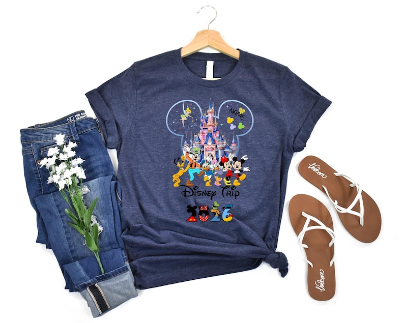 May include: A dark heather navy blue t-shirt featuring a graphic of a castle inside a Mickey Mouse ear silhouette, surrounded by cartoon characters, the text "Disney Trip", and the year "2026" with character-themed patterns on the numbers.