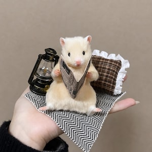 May include: A miniature cream-coloured hamster figurine holding a tiny newspaper. Beside the hamster is a small black and gold lantern and a brown plaid pillow with a white ruffled edge, all resting on a black and white chevron patterned fabric square.
