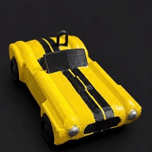 May include: A yellow toy car with black racing stripes down the center. The miniature vehicle has black wheels and silver headlights. The car is a classic roadster style, with a low profile and open cockpit. The background is black.