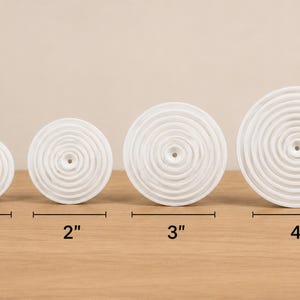 May include: Four white circular objects with concentric ripple patterns and a central hole. The objects are arranged in a row, with labels indicating diameters of 1 inch, 2 inches, 3 inches, and 4 inches respectively, measured by black lines.