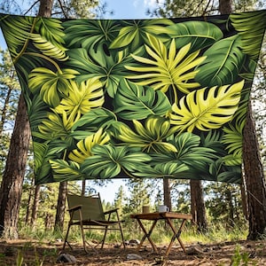 May include: A large, rectangular tapestry with a tropical leaf design in green and yellow against a black backdrop. The tapestry is suspended between trees, with a folding chair and small table set up beneath it. The scene is set outdoors.