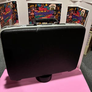 May include: A black rectangular hard-shell briefcase with a textured surface, featuring a silver-toned metal handle and matching silver-toned latches on the top edge. The case has a smooth, solid black front panel and a sturdy, structured frame.