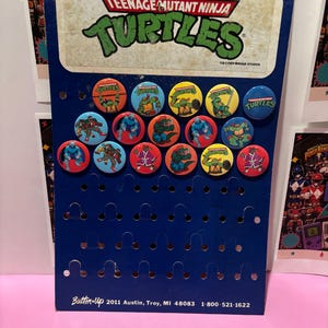 May include: A blue cardboard display board featuring the Teenage Mutant Ninja Turtles logo at the top and fourteen circular pin-back buttons with various character illustrations. The bottom text reads Button-Up 2011 Austin, Troy, MI 48083 1-800-521-1622.