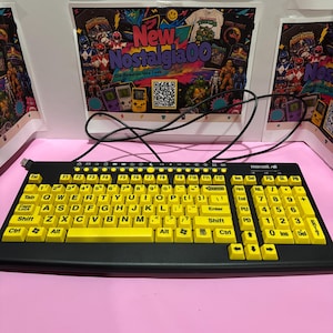 May include: A black Maxell computer keyboard featuring bright yellow keycaps with black lettering. The keyboard includes a full QWERTY layout, a numeric keypad, and a black USB cable extending from the top. The keys are arranged in a standard configuration with function keys across the top row and directional arrows near the numeric pad.