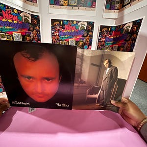Op de afbeelding: Een vinyl platenhoes voor Phil Collins getiteld No Jacket Required. De linkerkant bevat een close-up portret van Phil Collins met rode verlichting, terwijl de rechterkant een afbeelding van hem in een pak laat zien terwijl hij in een kamer staat.