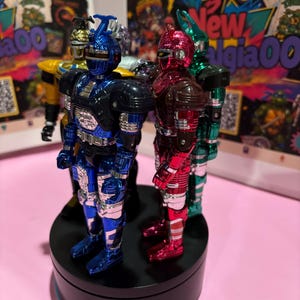 May include: A group of four metallic action figures standing on a black circular rotating display base. The figures feature shiny blue, red, green, and yellow finishes with insect-like helmet designs and detailed armor plating on their bodies.