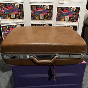 May include: A rectangular brown suitcase with a textured faux leather finish, featuring a silver-toned metal frame, two locking latches, and a central handle. A small brown luggage tag hangs from the handle of this vintage-style hard-shell case.