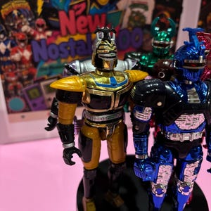 May include: Two metallic action figures standing on a black circular base. The figure on the left is gold and black with a horned helmet. The figure on the right is blue and black with a silver chest plate and a prominent horn on its helmet.