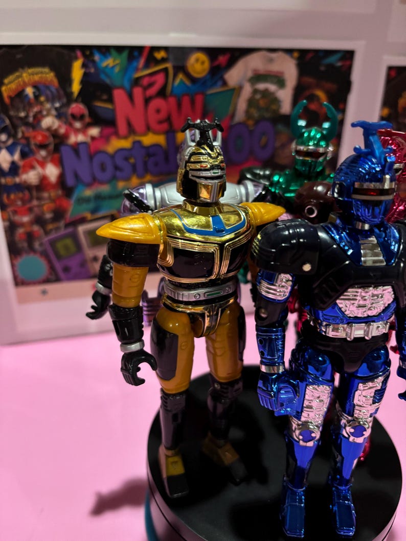 May include: Two metallic action figures standing on a black circular base. The figure on the left is gold and black with a horned helmet. The figure on the right is blue and black with a silver chest plate and a prominent horn on its helmet.