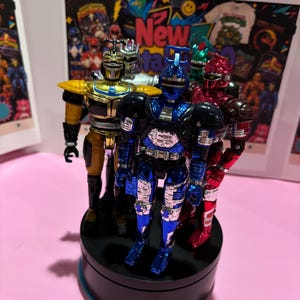May include: A group of five metallic action figures standing on a black circular display base. The figures feature shiny finishes in blue, gold, silver, red, and green, each with distinct insect-like head designs and articulated armor plating.