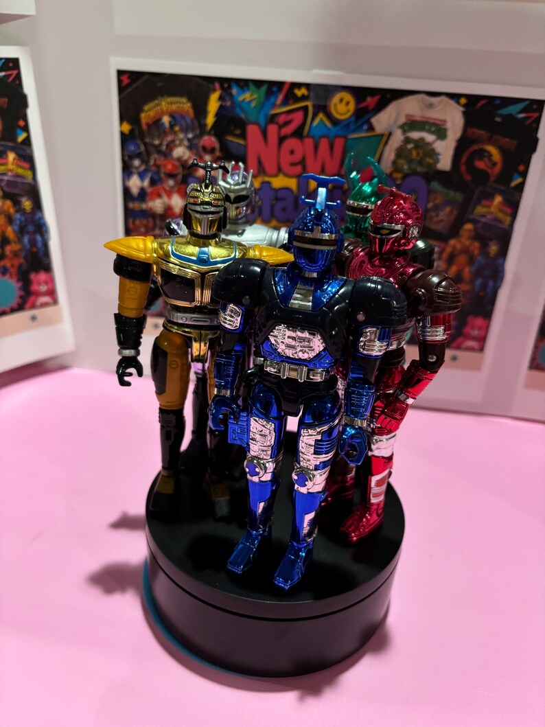 May include: A group of five metallic action figures standing on a black circular display base. The figures feature shiny finishes in blue, gold, silver, red, and green, each with distinct insect-like head designs and articulated armor plating.