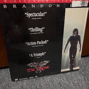 May include: A widescreen laserdisc of the film The Crow starring Brandon Lee. The black cover features white text quotes from critics, a graphic of a crow, and a vertical image of a figure in black clothing standing in a light-filled doorway.