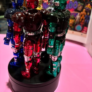 May include: A group of five metallic action figures with glossy finishes in blue, red, dark red, silver, and green. Each figure features a distinct helmet design with horn-like protrusions, standing upright on a circular black rotating display base.