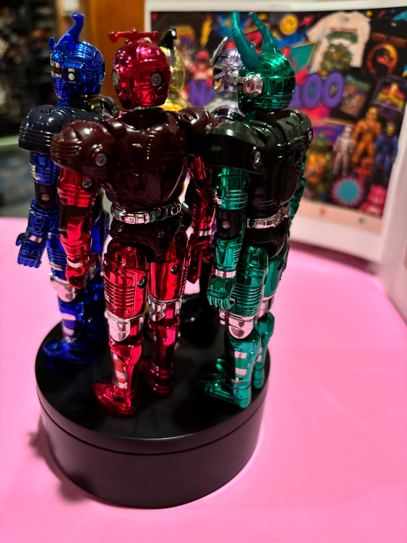 May include: A group of five metallic action figures with glossy finishes in blue, red, dark red, silver, and green. Each figure features a distinct helmet design with horn-like protrusions, standing upright on a circular black rotating display base.