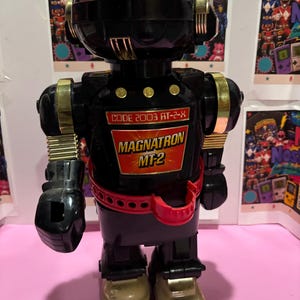 Może przedstawiać: Czarny robot zabawka ze złotymi akcentami, czerwoną półprzezroczystą kopułą na głowie i czerwonym paskiem. Na klatce piersiowej znajduje się etykieta z napisem "CODE 2003 AT-2-X" oraz czerwono-żółty emblemat "MAGNATRON MT-2" z beżowymi stopami.