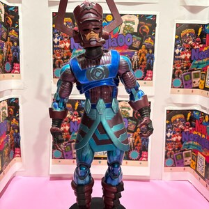 Può includere: Un action figure di Galactus con un alto elmo viola cornuto, un'armatura blu metallizzato e viola scuro con un emblema circolare sul petto e arti articolati, posizionato su una base espositiva nera.