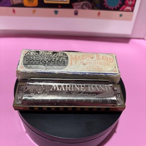Könnte beinhalten: Eine silberfarbene M. Hohner Marine Band Mundharmonika mit zehn nummerierten Löchern und passender Pappschachtel. Die Mundharmonika trägt die Gravur Marine Band M. Hohner sowie den Buchstaben G. Die Schachtel zeigt ein altes Schwarz-Weiß-Foto.