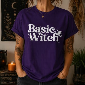 May include: A deep purple short-sleeved t-shirt featuring the white text "Basic Witch" printed on the front. The design includes small star icons and a stylised witch hat graphic integrated into the lettering. The shirt has a crew neckline.