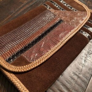 Può includere: Una pochette in camoscio marrone con chiusura a patta. La patta presenta un design patchwork con pannelli strutturati e fantasia nei toni del marrone, nero e beige. I bordi sono rifiniti con un bordo cucito marrone chiaro.