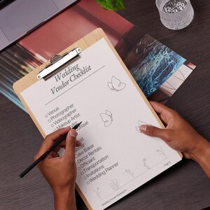 K&ouml;nnte beinhalten: Ein helles Holz-Klemmbrett mit einem wei&szlig;en Blatt Papier, das mit Wedding Vendor Checklist betitelt ist. Das Papier enth&auml;lt eine Liste von Hochzeitsdienstleistungen mit Kontrollk&auml;stchen, Schmetterlingszeichnungen und kleinen Blumen.