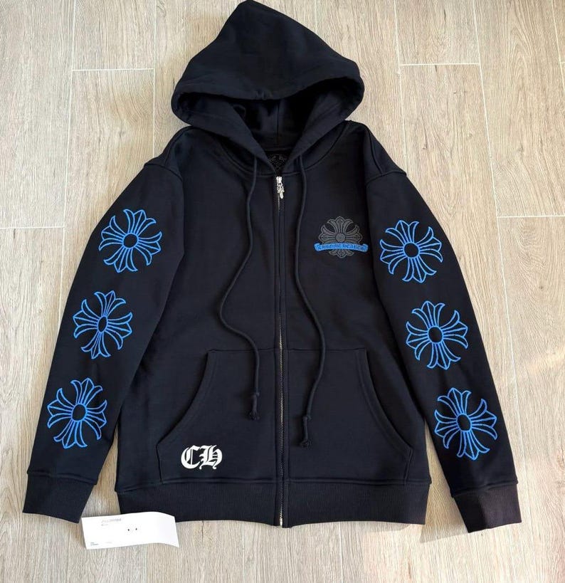 Puede incluir: Sudadera negra con cremallera, capucha y bolsillos delanteros. Las mangas presentan dise&ntilde;os florales azules y el pecho un emblema similar. La sudadera tiene una cremallera plateada y las letras "CH" en blanco.