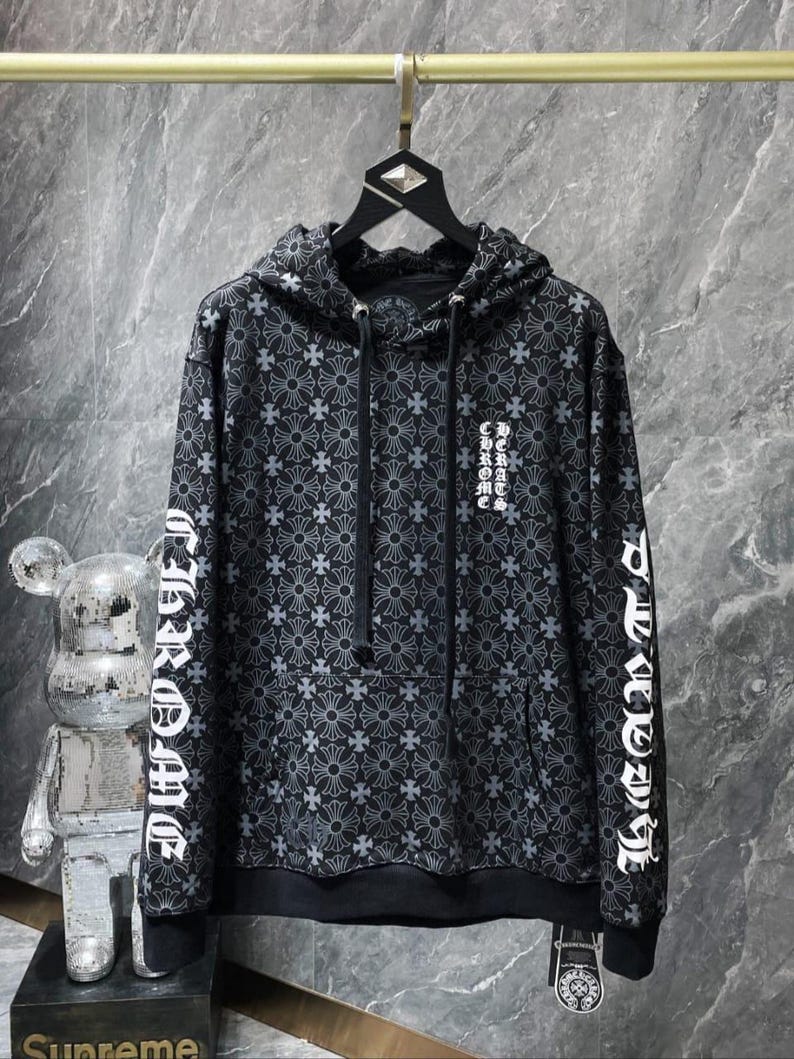 May include: Black hoodie with a repeating cross pattern and white text on the sleeves and chest. The hoodie has a drawstring hood and a front pocket. The garment is hanging on a gold-colored rod.