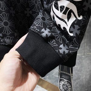 May include: Black hoodie with a floral pattern and white lettering on the sleeve. The hoodie has a black ribbed cuff and a tag with a logo. The floral pattern is a repeating design in shades of gray.