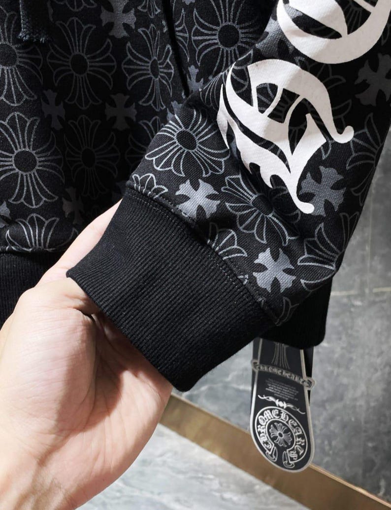May include: Black hoodie with a floral pattern and white lettering on the sleeve. The hoodie has a black ribbed cuff and a tag with a logo. The floral pattern is a repeating design in shades of gray.