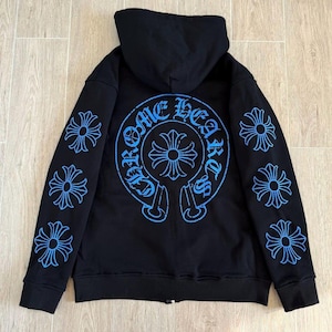 Puede incluir: Sudadera con capucha negra con cremallera, capucha y dise&ntilde;os gr&aacute;ficos azules. La parte trasera presenta un gran dise&ntilde;o circular con las palabras "Chrome Hearts" y una cruz. Las mangas tienen m&uacute;ltiples dise&ntilde;os en forma de cruz.
