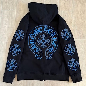 Puede incluir: Sudadera con capucha negra con cremallera y dise&ntilde;os gr&aacute;ficos azules. La parte trasera presenta un gran dise&ntilde;o circular con el texto "Chrome Hearts" y un emblema de cruz. Las mangas tienen m&uacute;ltiples dise&ntilde;os florales azules en forma de cruz.