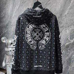 May include: Black hoodie with a repeating floral pattern and white accents. The back features a large white emblem with text, and the sleeves have white text. The hoodie is hanging on a metal rack.