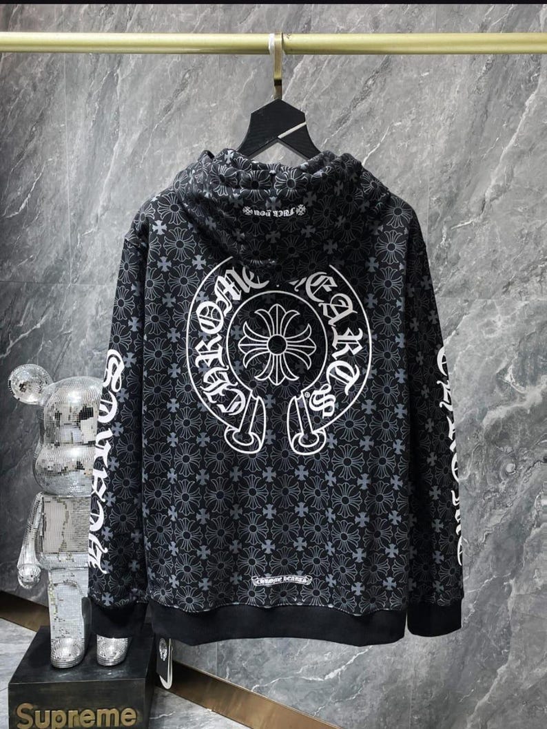 May include: Black hoodie with a repeating floral pattern and white accents. The back features a large white emblem with text, and the sleeves have white text. The hoodie is hanging on a metal rack.