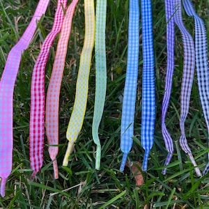 May include: A collection of nine flat shoelaces featuring a gingham check pattern in a variety of colors including pink, red, orange, yellow, light green, light blue, dark blue, purple, and black, each with a solid colored plastic aglet tip.