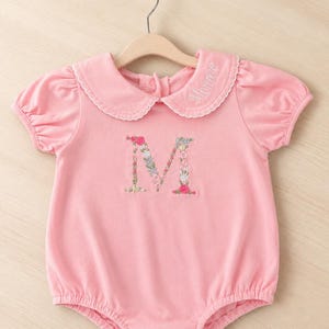 May include: Pale pink baby romper with a Peter Pan collar and puff sleeves. The collar is trimmed with white lace and has the name "Monroe" embroidered. The letter "M" is embroidered on the front with floral detailing.