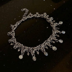May include: A silver-toned chainmaille anklet featuring a complex woven link pattern. The piece is adorned with small dangling metal heart charms and translucent, pale, round beads suspended from the main chain, finished with a lobster clasp.