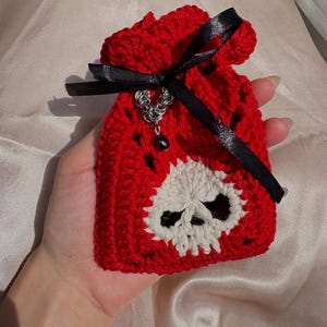 Pode incluir: Uma pequena bolsa de crochê vermelha com cordão e um motivo de caveira branca na frente. A bolsa é fechada com uma fita de cetim preta e decorada com um pingente de cota de malha prateada que inclui uma pequena conta preta pendurada.