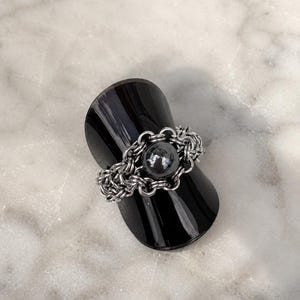 May include: A silver-toned chainmail ring featuring a central dark gray spherical bead. The ring is constructed from multiple interlocking metal jump rings that form a textured band, with the smooth, round bead secured in the center of the design.