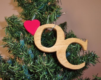 May include: Wooden Christmas ornament in the shape of the letters "OC" with a red heart. The ornament is hanging on a green Christmas tree with small white lights.