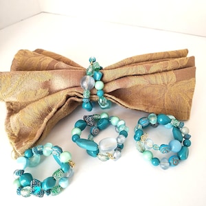 May include: A set of four beaded napkin rings in shades of turquoise, teal, and clear. The beads are various shapes and sizes, with some featuring patterned designs. A gold-colored napkin is folded in the background.